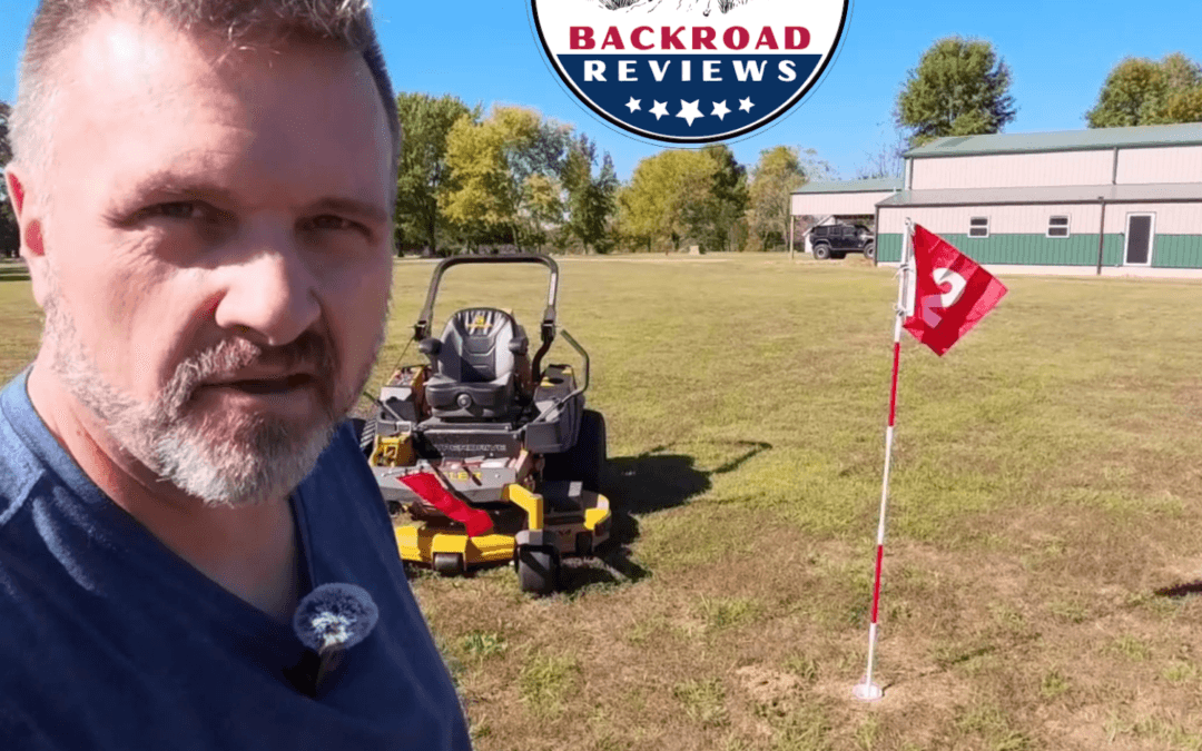 This is a bright outdoor selfie-style photo taken on a sunny day in a large, open grassy field (a mowed yard or pasture). In the foreground on the left, a middle-aged man with short, graying brown hair and a salt-and-pepper beard looks directly at the camera. He is wearing a blue t-shirt and has a small lavalier microphone clipped to his collar. Behind him sits a yellow zero-turn riding lawn mower. To the right of the mower stands a red golf flag numbered “12” on a white-and-red striped flagstick that is planted in a white cup in the ground. In the background is a large green-and-beige metal building (barn/workshop style) with a dark vehicle parked nearby, along with trees showing some autumn foliage under a clear blue sky. A circular “BACKROAD REVIEWS” logo (“Quality Reviews & Honest Opinions”) is overlaid at the top center of the image.