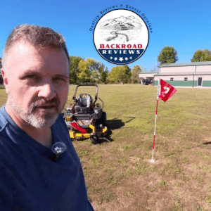 Golf Flagstick 6ft Golf Flag and Cup for Yard Pro Detachable Golf Hole Cup and Flag for Driving Range Backyard Upgrade Anti-Rust Glass Fiber 5-Section Design with Connectors xl This is a bright outdoor selfie-style photo taken on a sunny day in a large, open grassy field (a mowed yard or pasture). In the foreground on the left, a middle-aged man with short, graying brown hair and a salt-and-pepper beard looks directly at the camera. He is wearing a blue t-shirt and has a small lavalier microphone clipped to his collar. Behind him sits a yellow zero-turn riding lawn mower. To the right of the mower stands a red golf flag numbered “12” on a white-and-red striped flagstick that is planted in a white cup in the ground. In the background is a large green-and-beige metal building (barn/workshop style) with a dark vehicle parked nearby, along with trees showing some autumn foliage under a clear blue sky. A circular “BACKROAD REVIEWS” logo (“Quality Reviews & Honest Opinions”) is overlaid at the top center of the image.